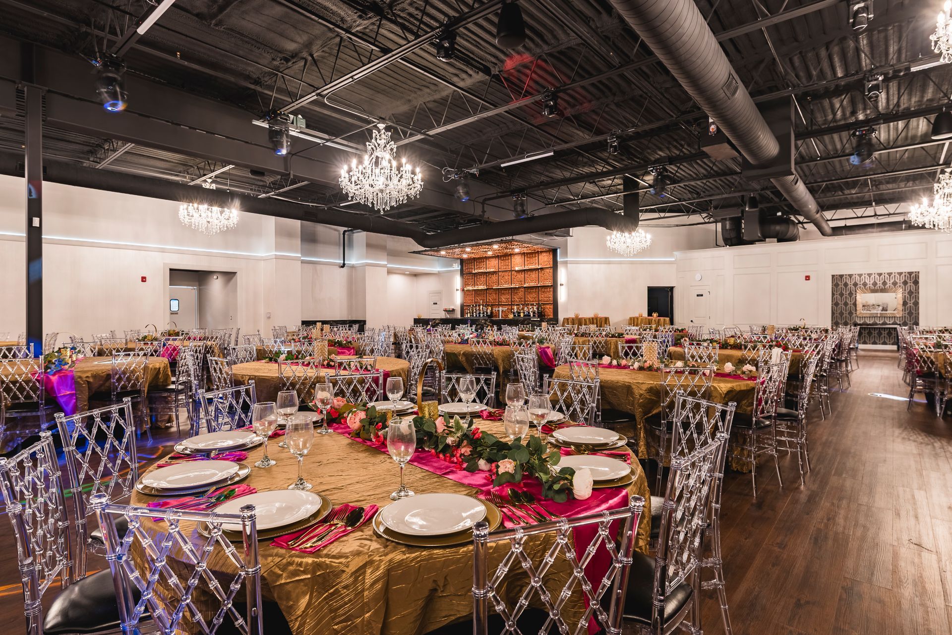 Decorated banquet hall with round tables, gold linens, clear chairs, and chandeliers under an industrial ceiling