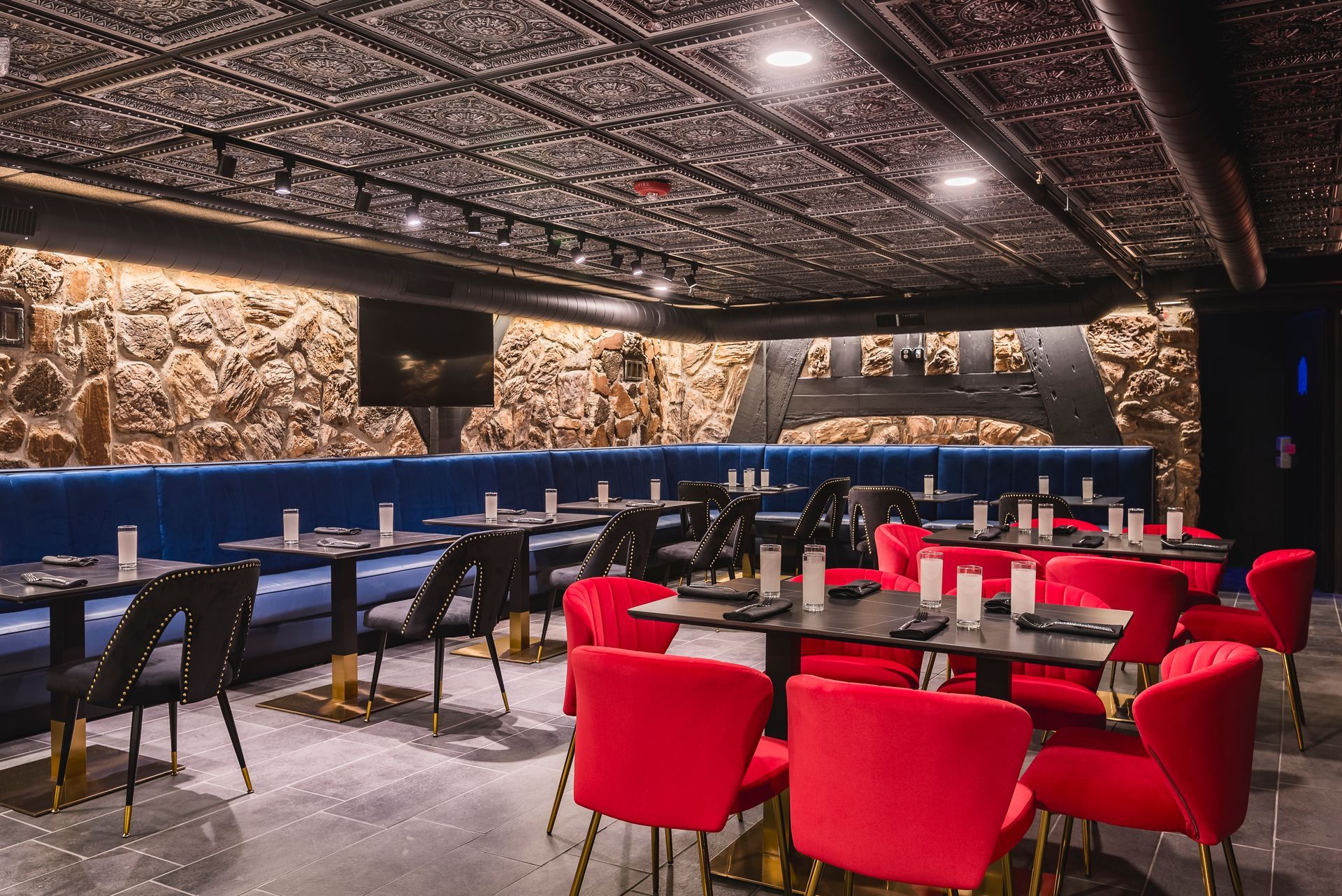 Modern lounge with red chairs, blue banquettes, and tables under a patterned ceiling in dim lighting