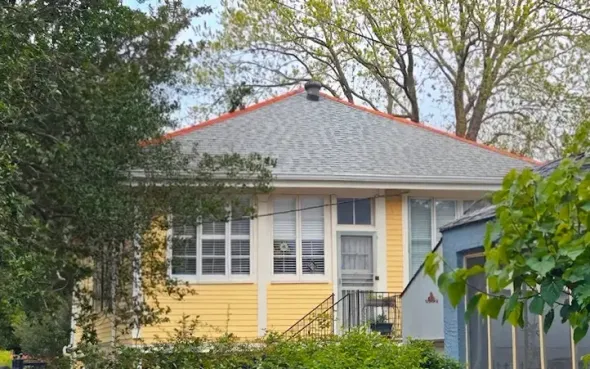 Yellow house with white trim, gray roof, and large windows, partly framed by trees and a blue shed