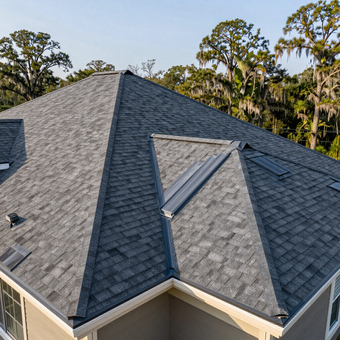 Aerial view of a gray shingle roof with a triangular skylight and trees in the background