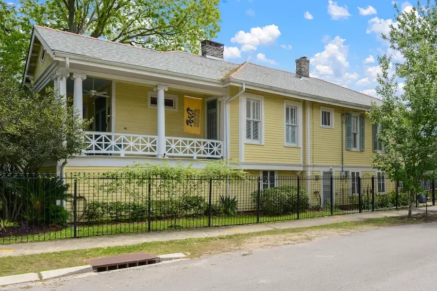 Yellow house with white trim and porch behind a black fence on a tree-lined street