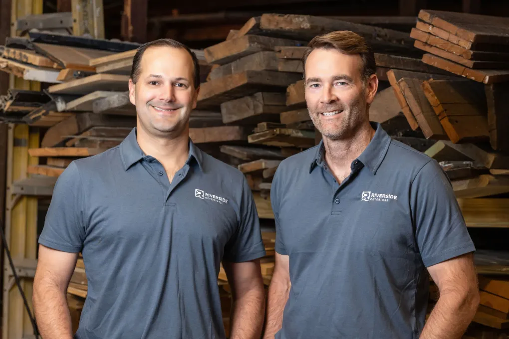 Owners in gray polos standing in a lumber warehouse, smiling in front of stacked wood.