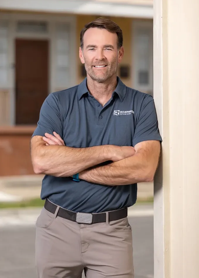 Man standing with arms crossed in a gray polo and khaki pants, smiling in a bright indoor hallway