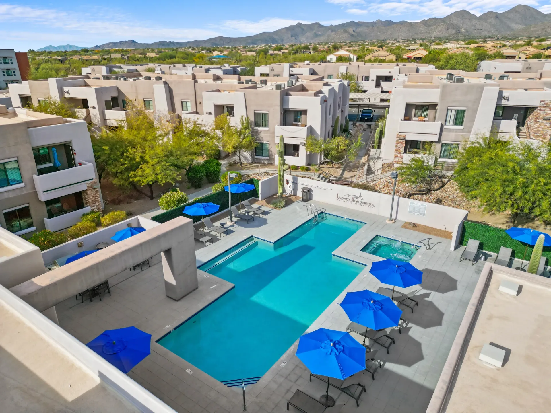 A community swimming pool and hot tub area with lounge chairs and blue umbrellas, surrounded by apartment buildings and desert landscaping.