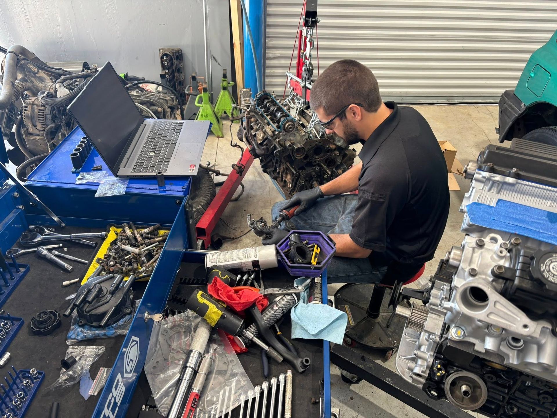 Mechanic working on an engine in a cluttered auto shop, with tools, laptop, and car parts nearby