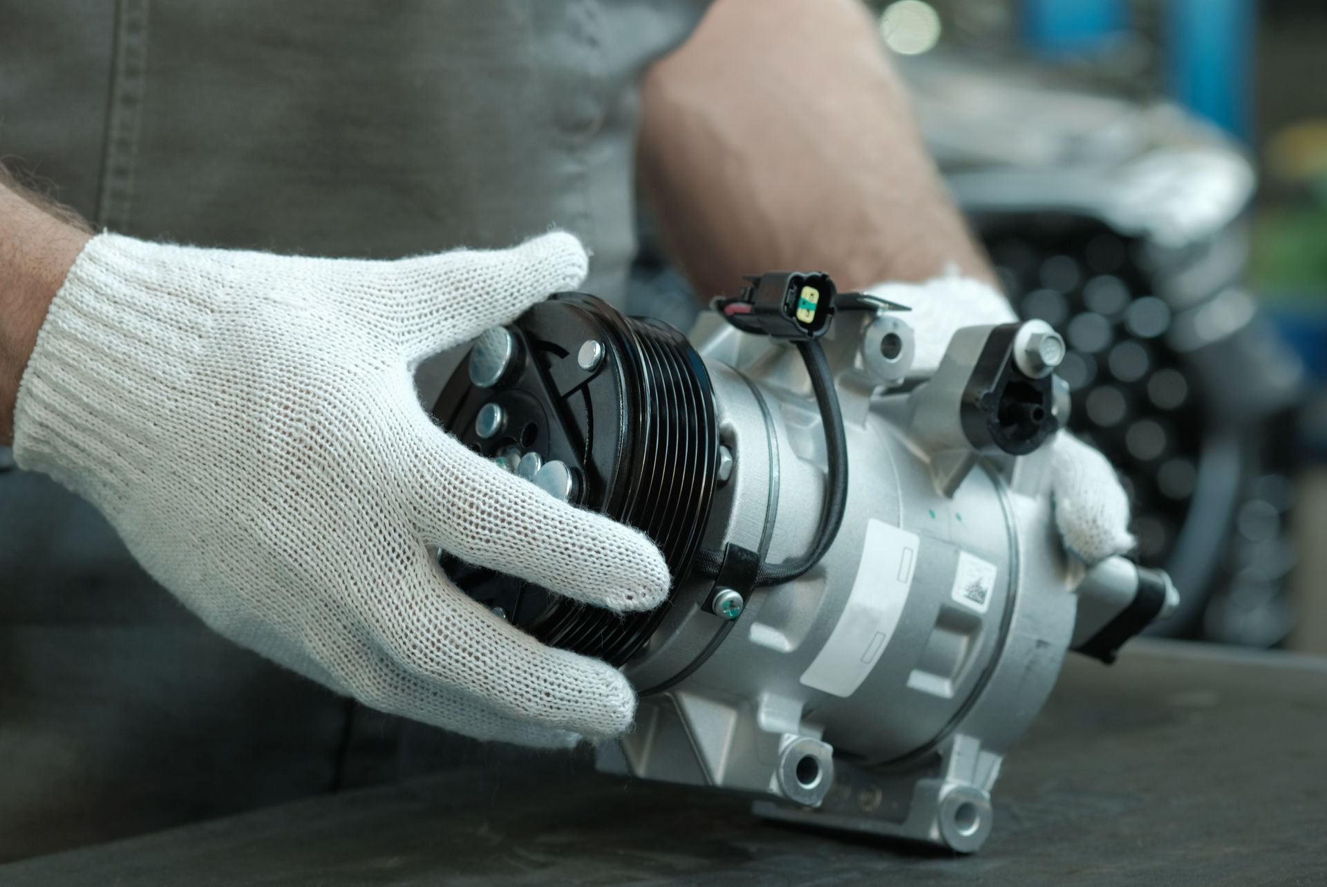 Gloved hands holding a small silver mechanical device on a workbench