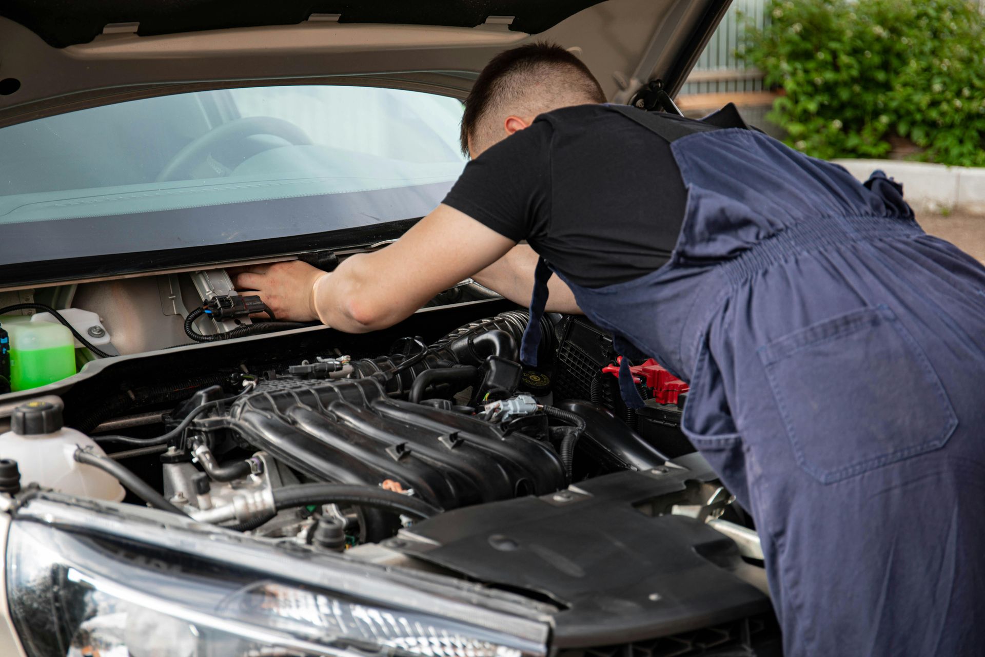 Mechanic in blue overalls working under an open car hood in a garage