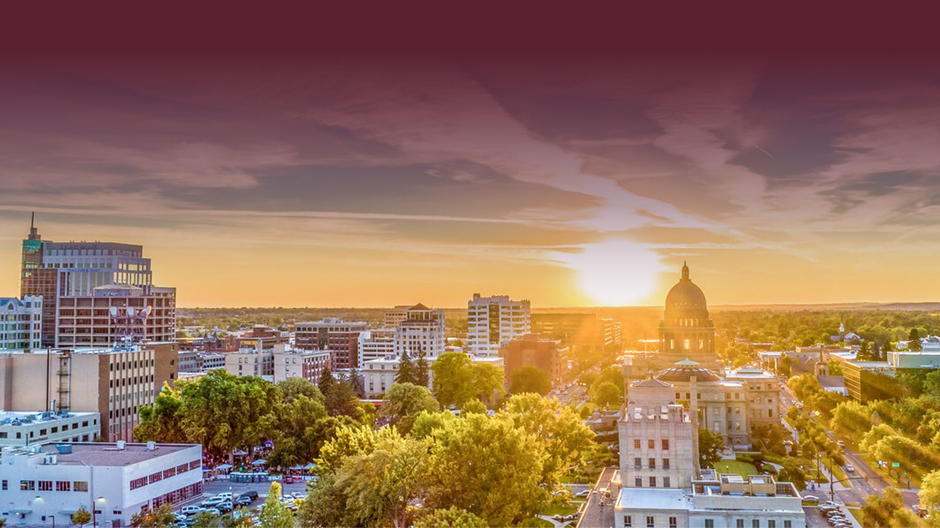 Sunset over a city skyline with trees, modern buildings, and a domed landmark in warm golden light