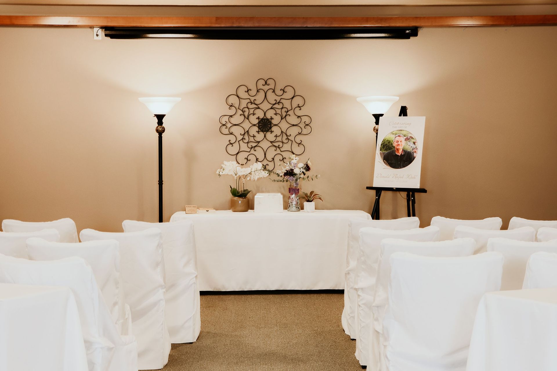 White ceremony seating facing a decorated table and floral wall display in a beige event room