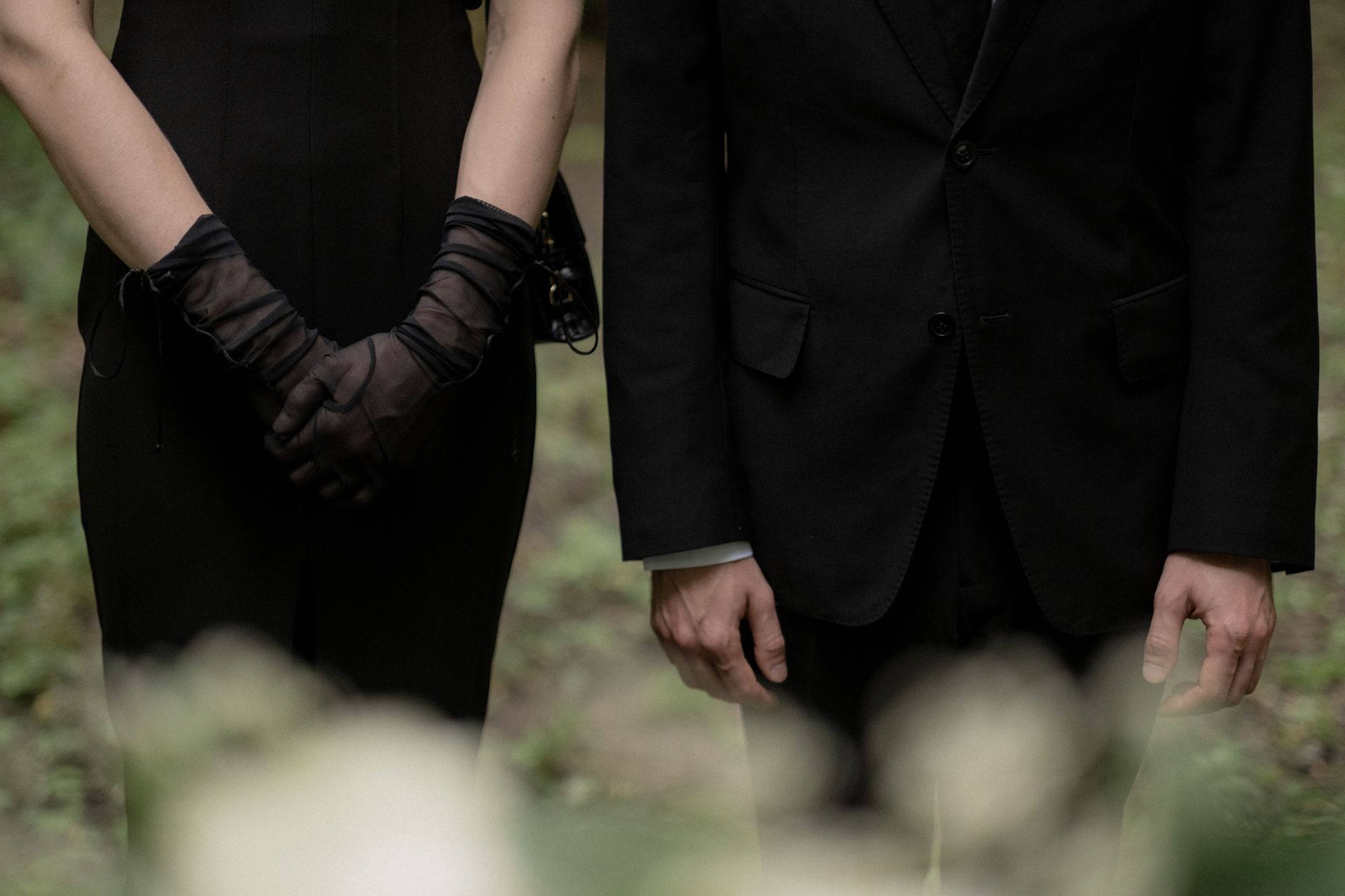 Two people in black formalwear stand with hands clasped, facing forward outdoors.