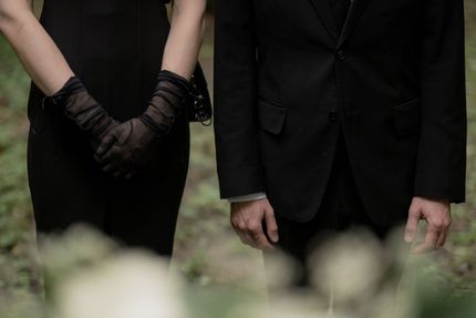 Two people in black formalwear stand with hands clasped, facing forward outdoors.