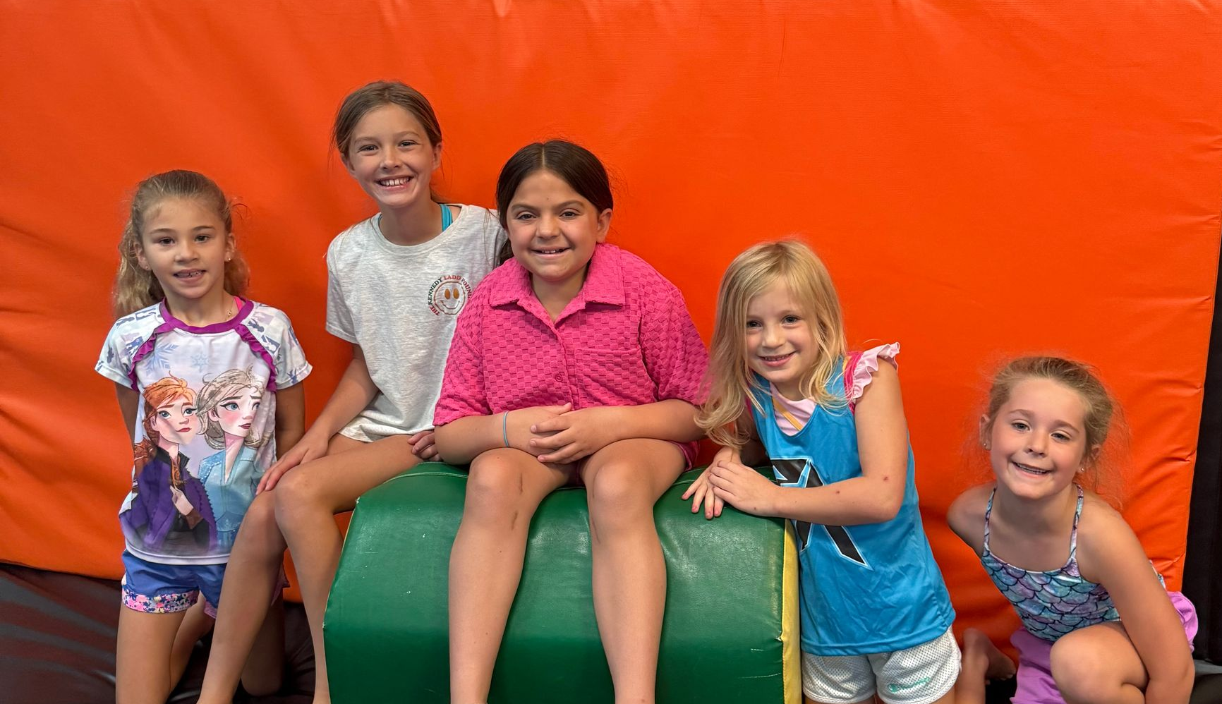 Five children smiling while posing together against an orange wall in an indoor activity space.