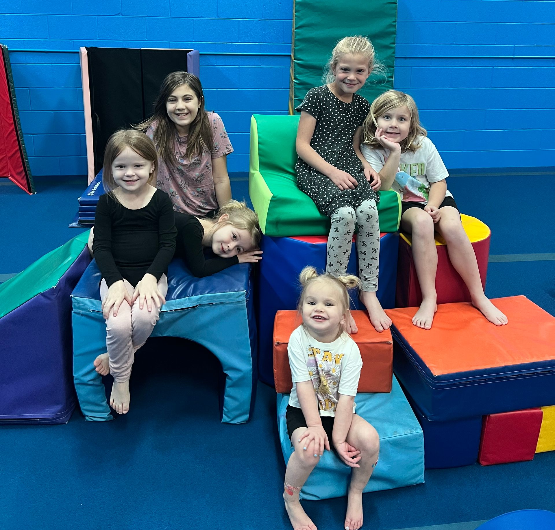 Five children sit on colorful gymnastic foam mats and blocks in a gym.