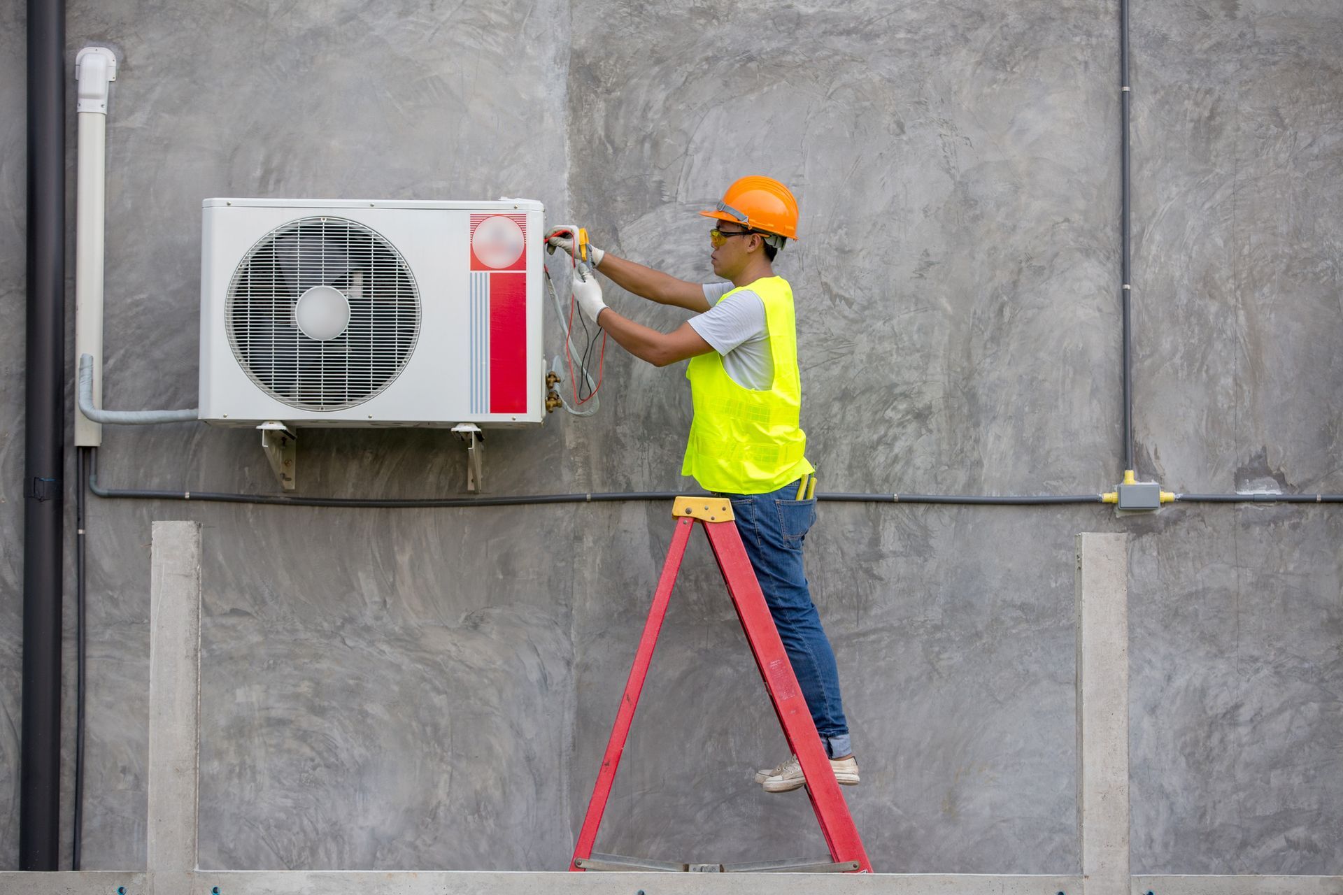 A person in a hard hat and safety vest is installing an air conditioner on a concrete wall, standing on a red ladder.