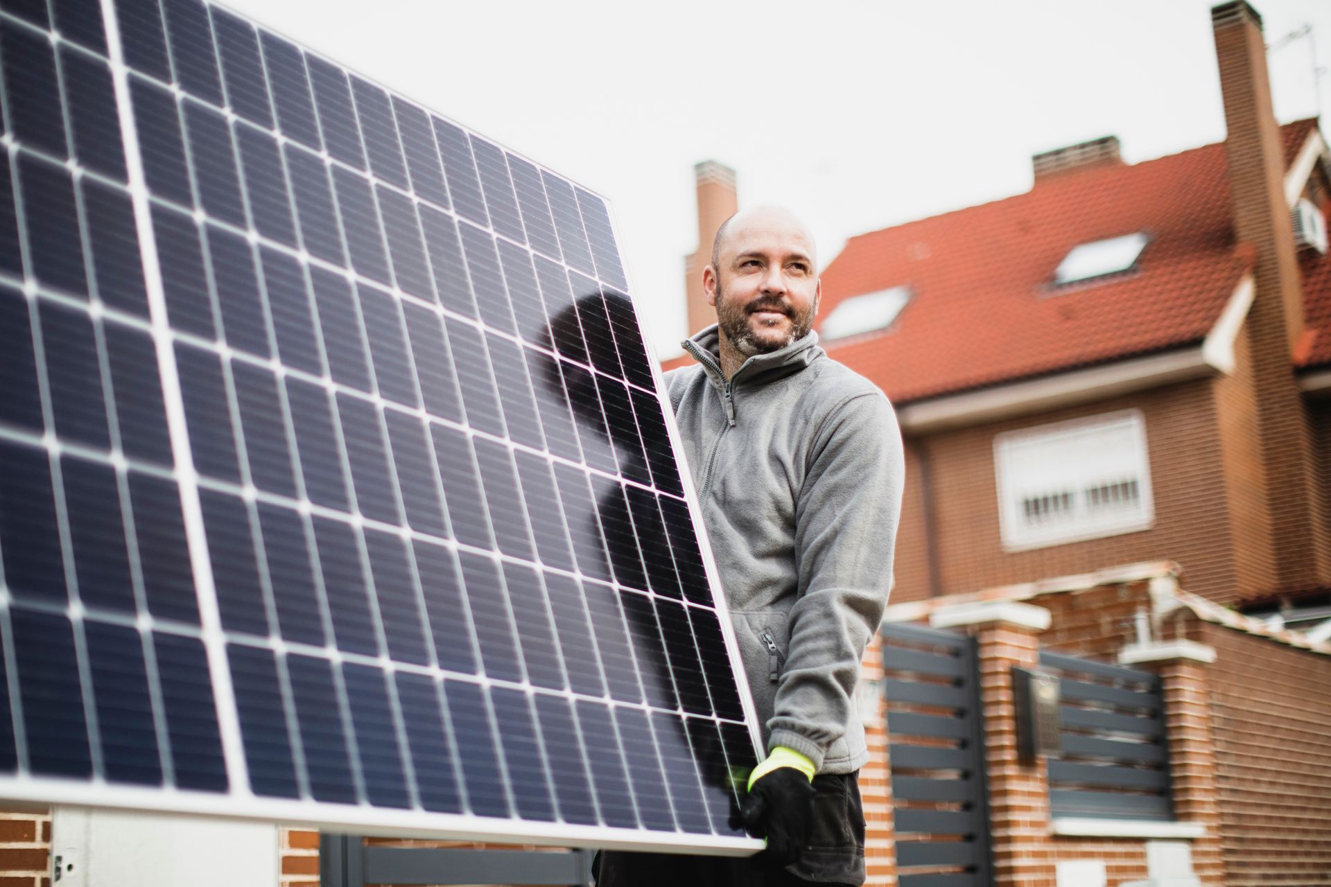 Man carrying a large solar panel towards a residential building; he smiles and appears to be installing it outside.