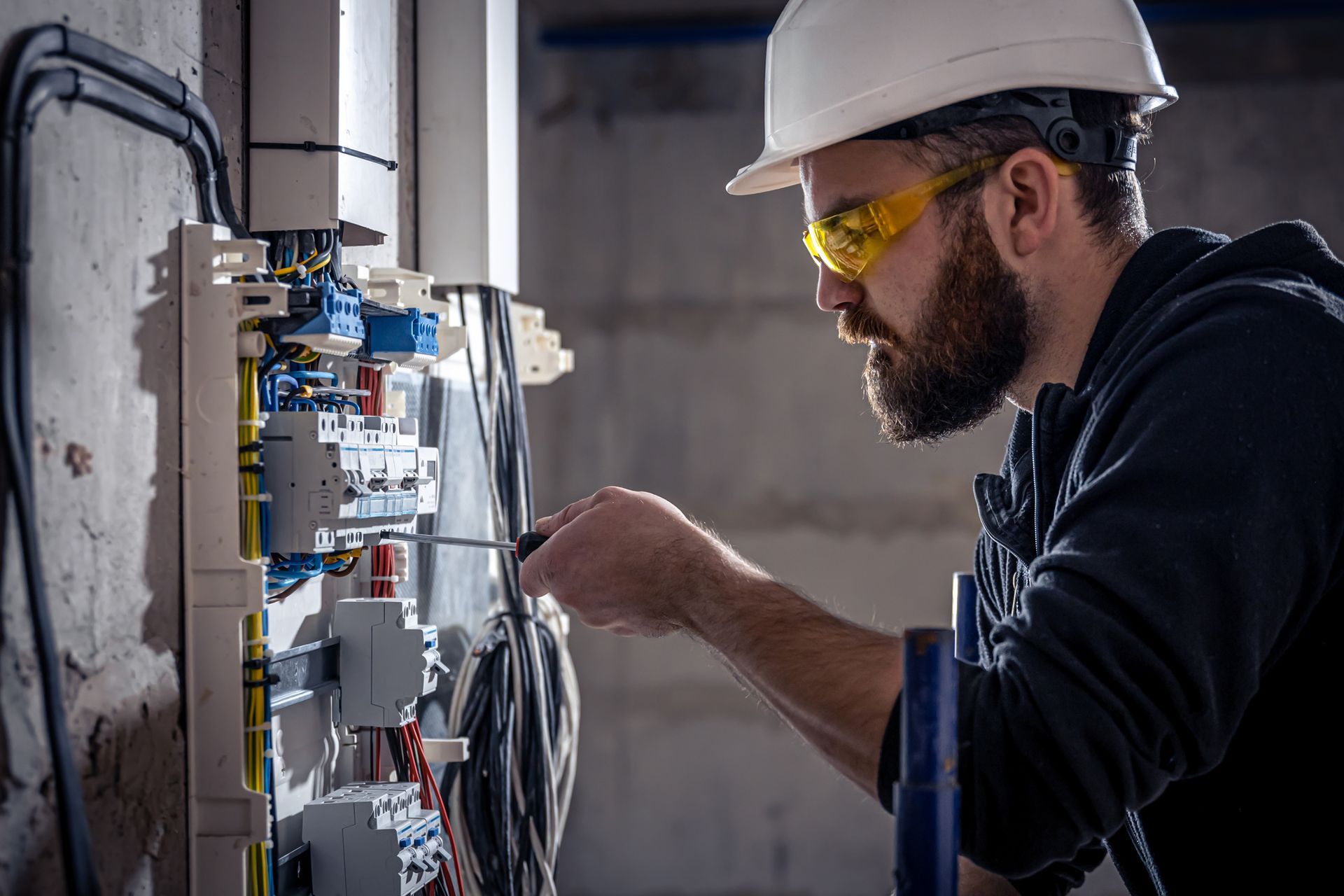 Electrician in a hard hat and safety glasses working on electrical panel.