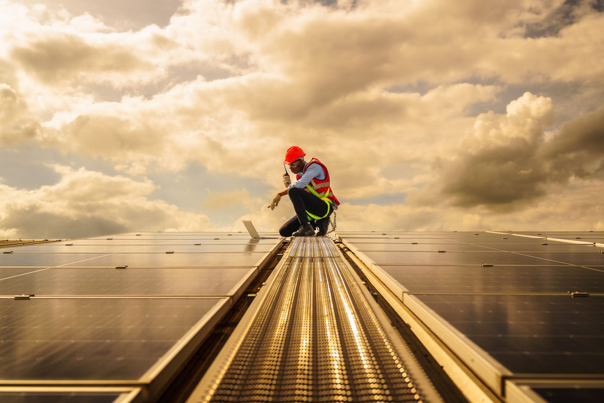 Man in safety gear inspecting solar panels on a rooftop, using a laptop, under a cloudy sky.