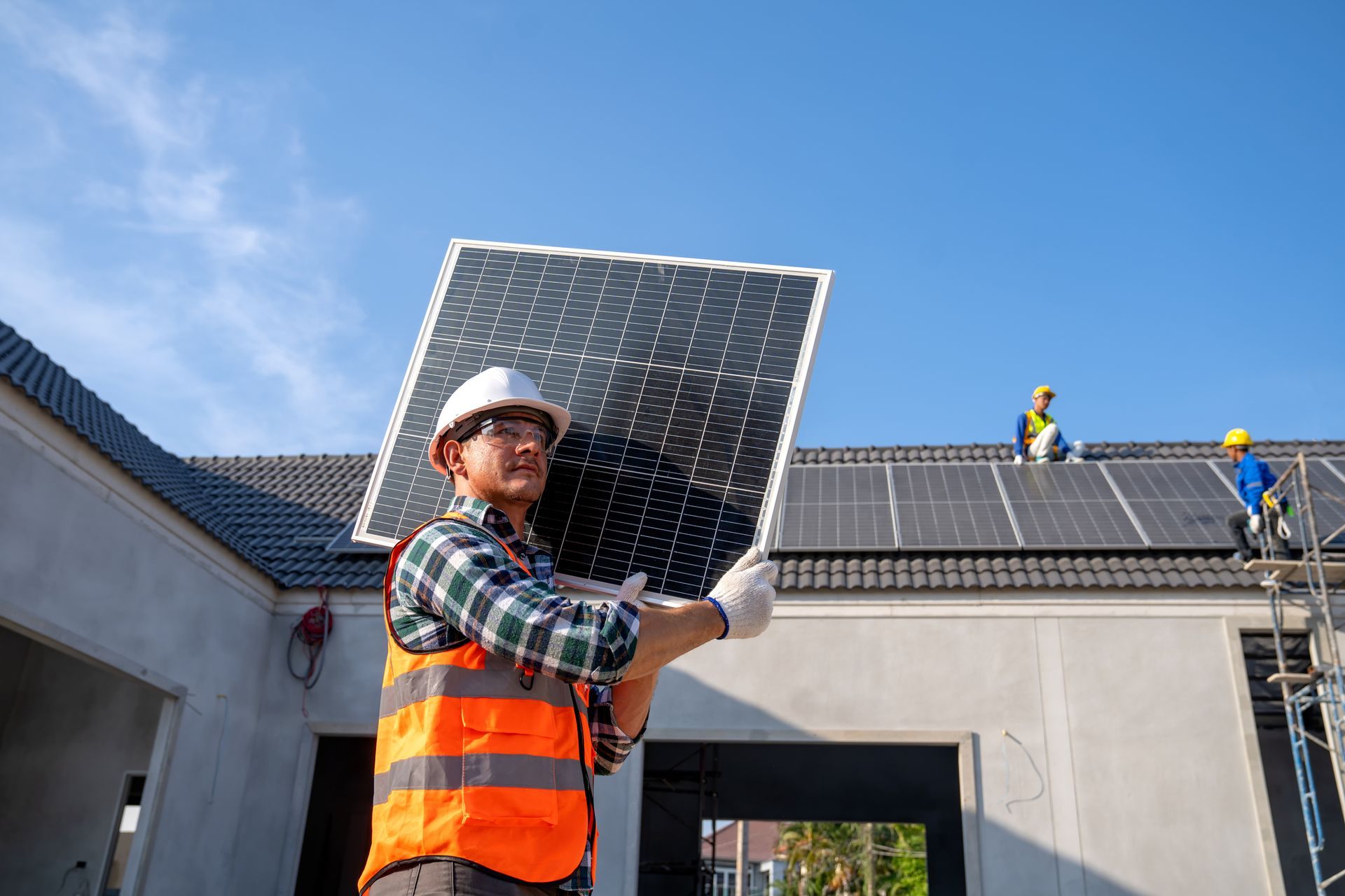 Construction worker carrying a solar panel on a rooftop under a clear blue sky. Other workers are installing solar panels on the roof.