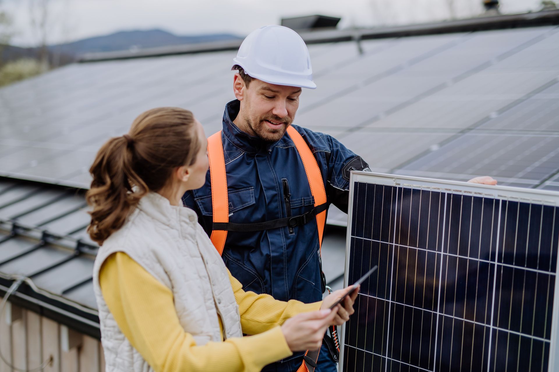 Woman and solar panel installer reviewing a tablet near a rooftop solar panel array. They are outdoors on a roof.