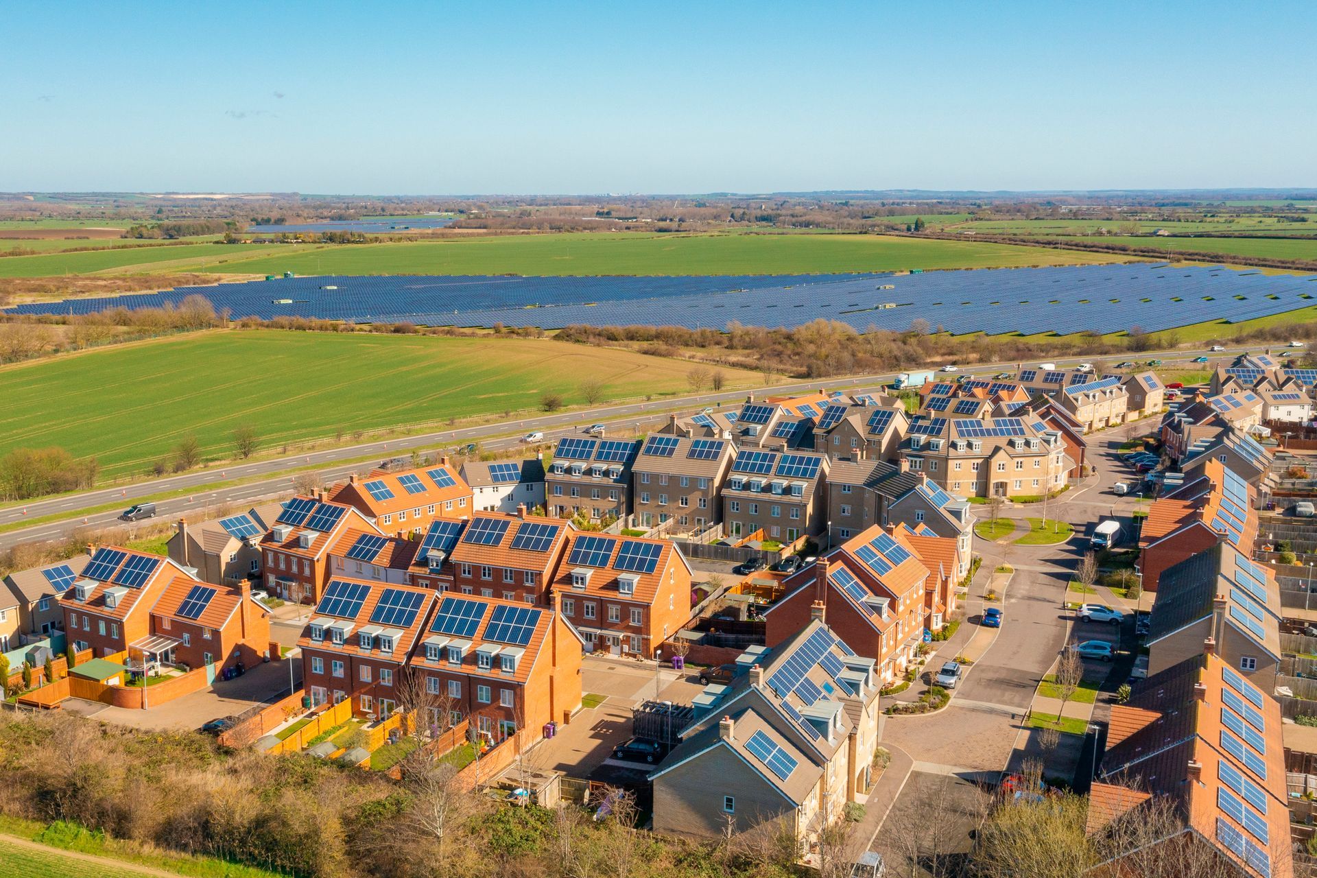 Aerial view of a residential neighborhood with solar panels on rooftops, adjacent to a large solar panel farm and green fields.
