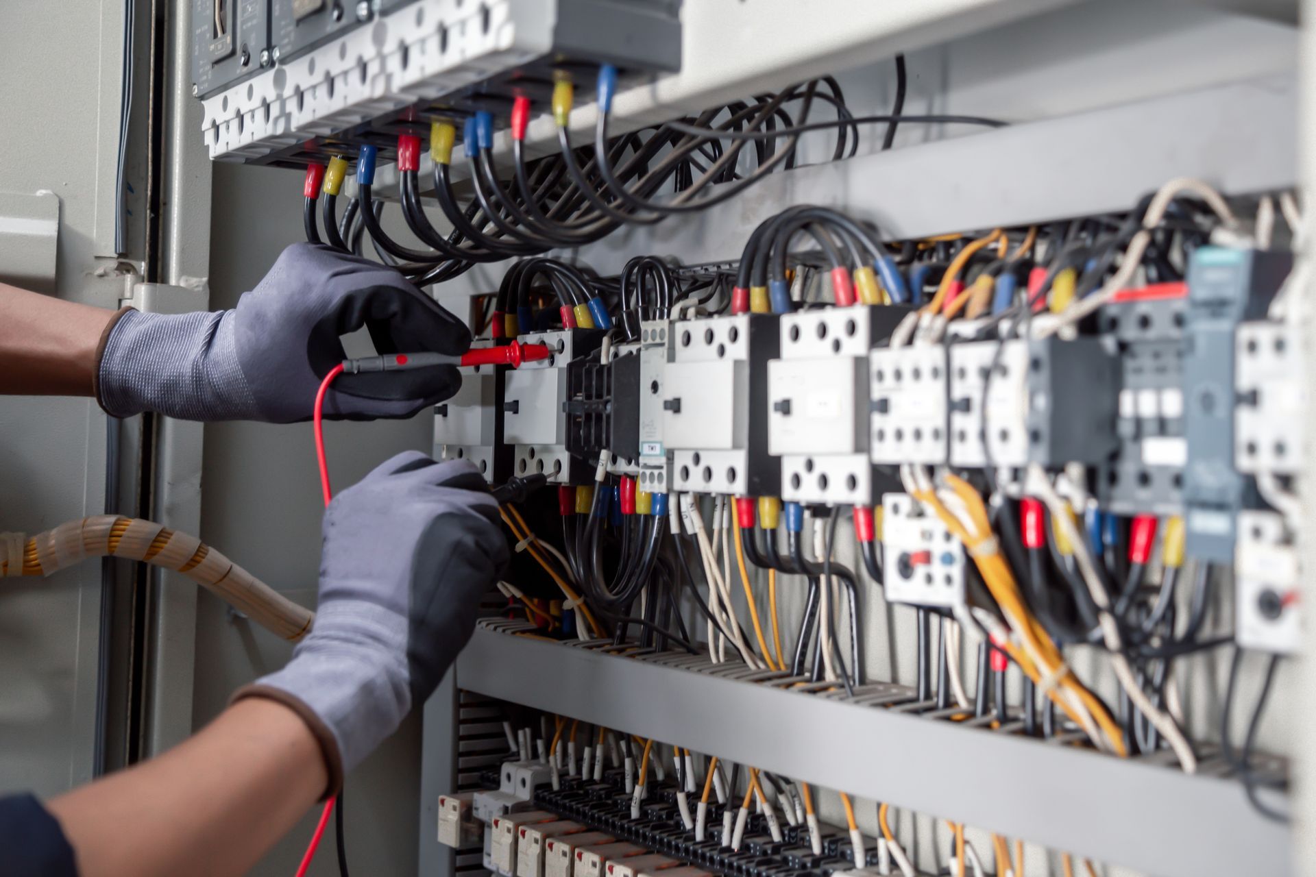 Hands wearing gloves using a multimeter to test wires in an electrical panel.