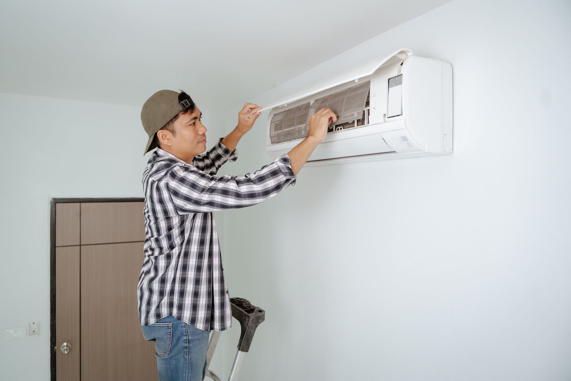 Man on a ladder cleans the filter of a wall-mounted air conditioner in a white room. He wears a plaid shirt and a cap.
