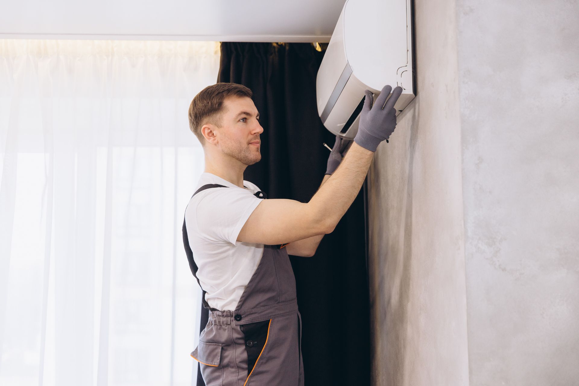 Man in grey overalls, wearing gloves, working on a wall-mounted air conditioner with a screwdriver. Indoors with curtains and a window.