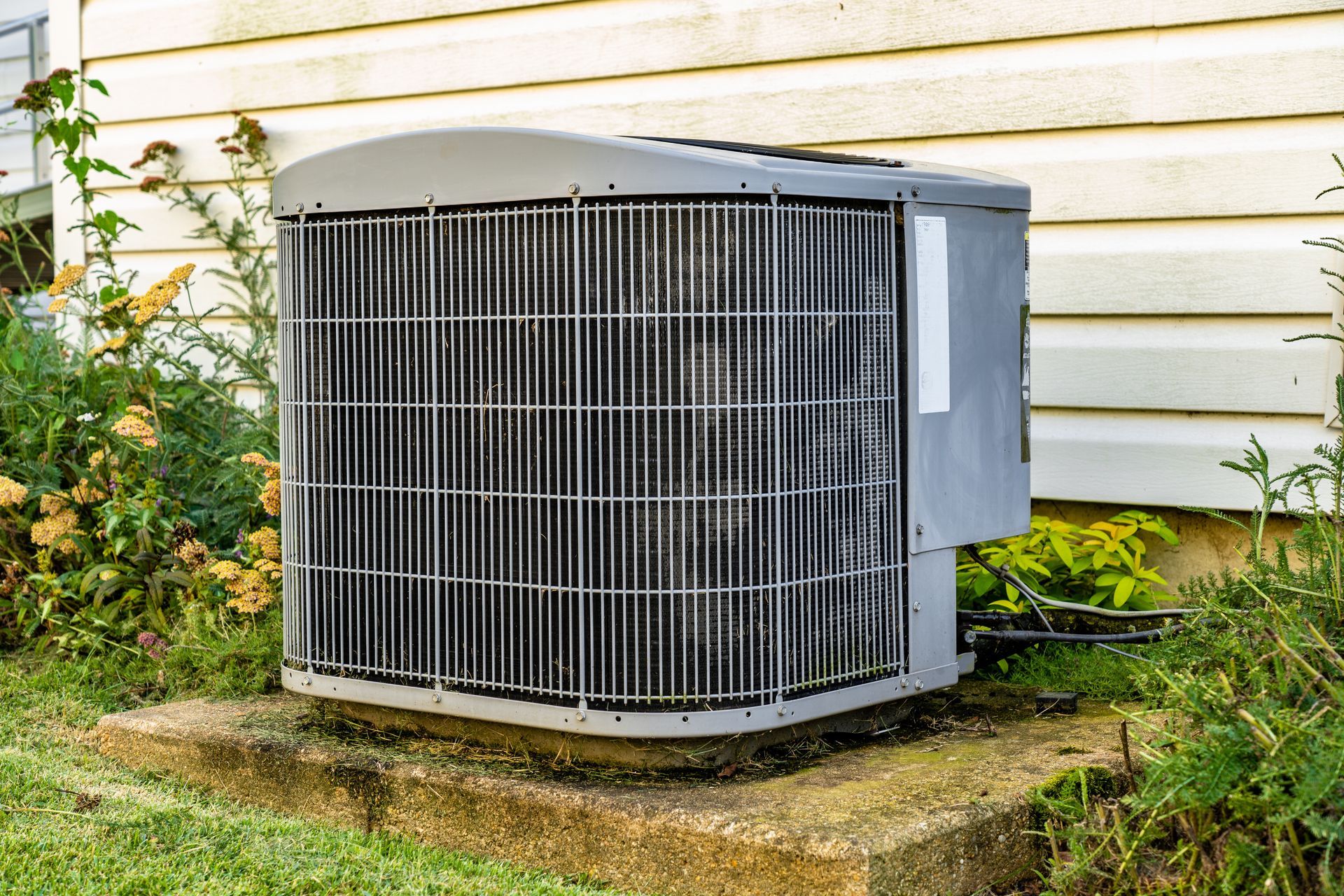 Outdoor air conditioner unit beside a house wall, with metal grille and gray casing