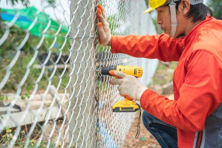 Construction worker fastening chain-link fence with a power drill outdoors.
