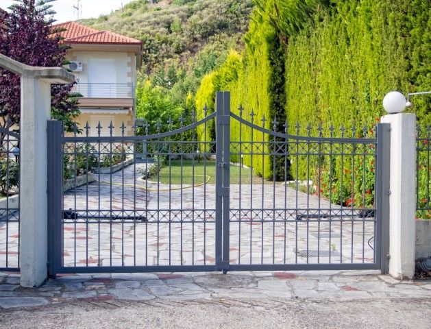 Dark gray metal gate, framed by white pillars, opens onto a driveway, with a house and green foliage in the background.