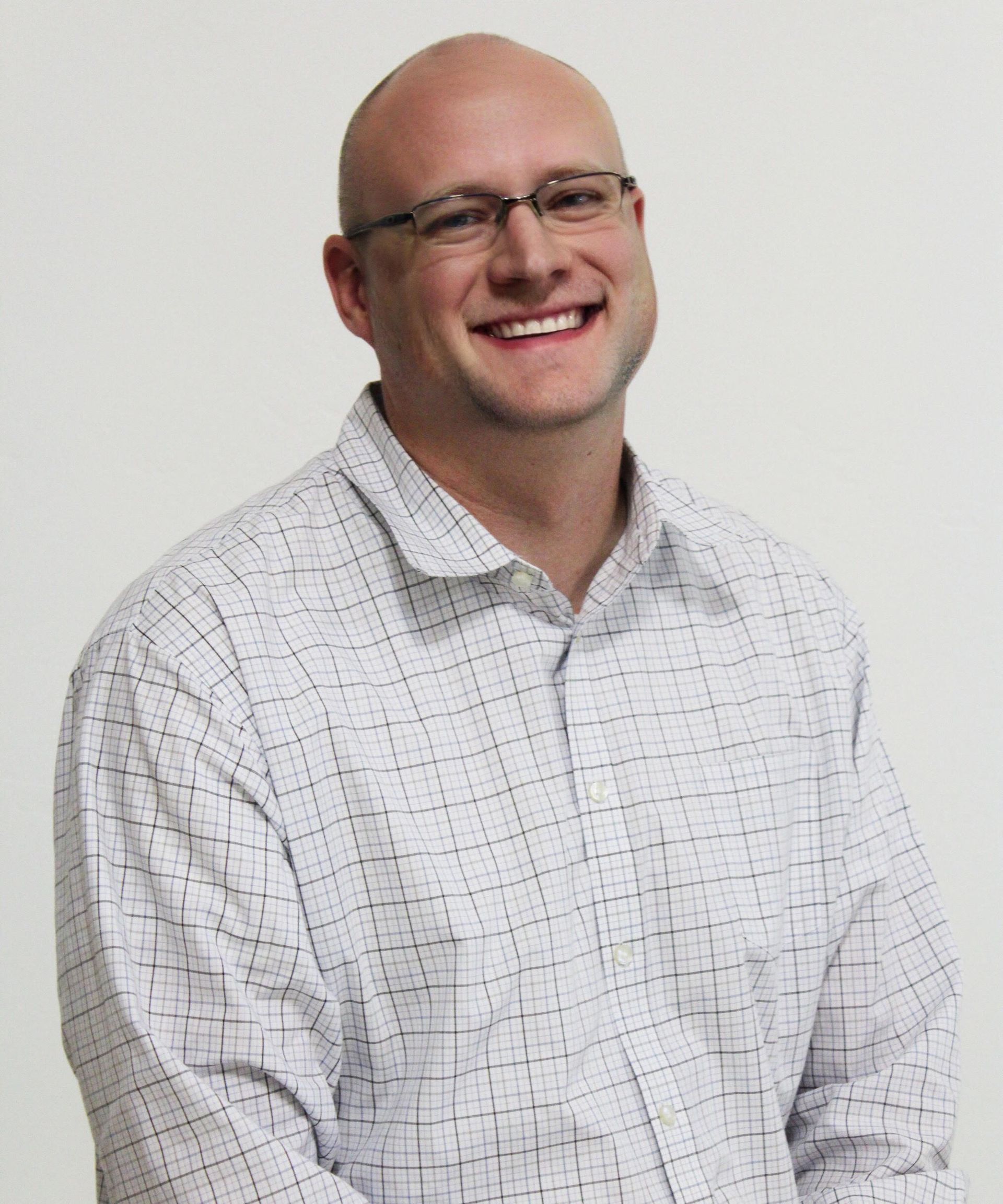 Man with glasses smiling, wearing a light-colored button-down shirt, set against a white backdrop.