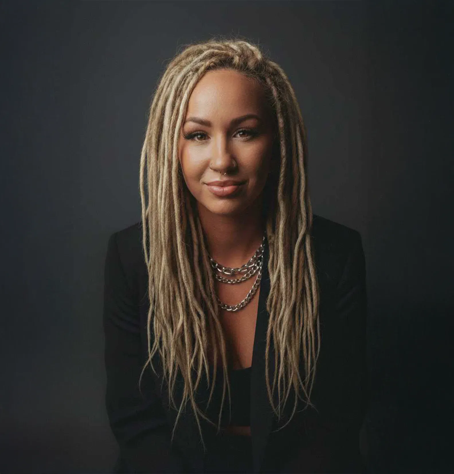 Woman with blonde dreadlocks, wearing a blazer and necklace, smiling against a dark background.