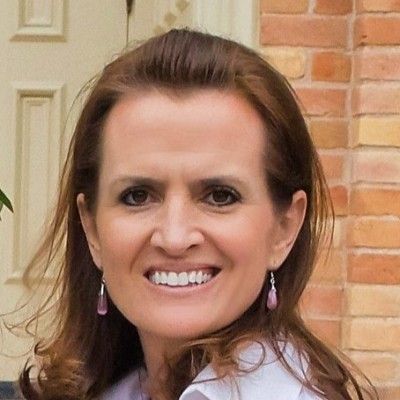 Smiling woman with brown hair wearing pink earrings, standing by a brick wall outdoors