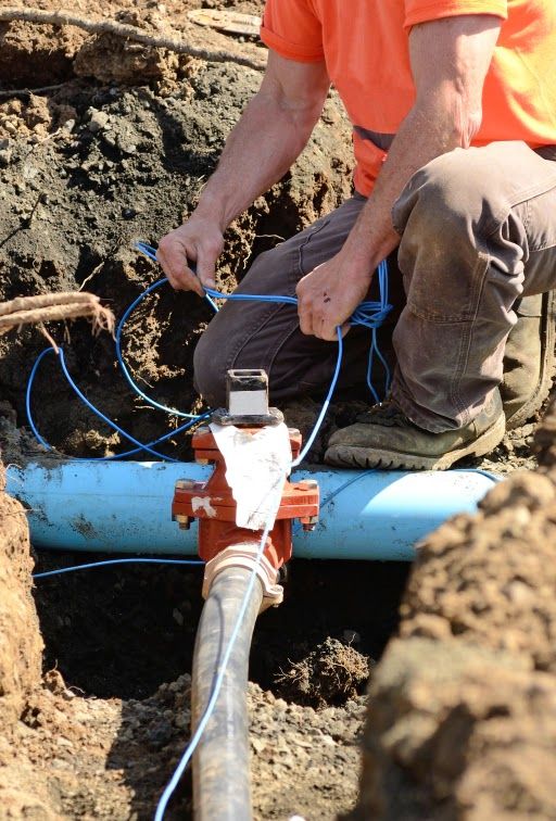 Man connecting blue wires to a water valve in a ditch, wearing an orange shirt and work boots.