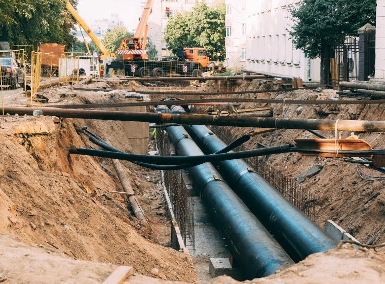 Construction site: Workers in a trench installing large black pipes; crane and buildings in the background.