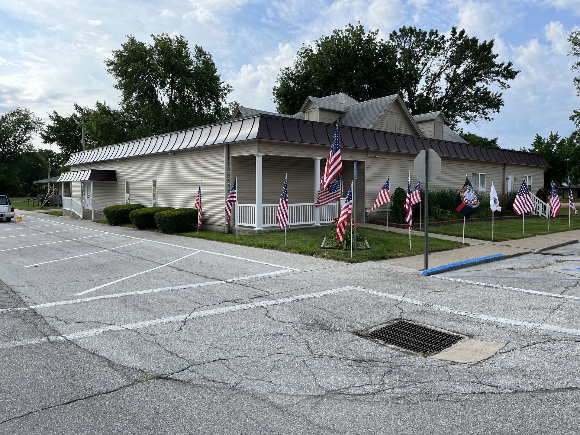Small beige building with multiple American flags along a roadside, under a cloudy sky.