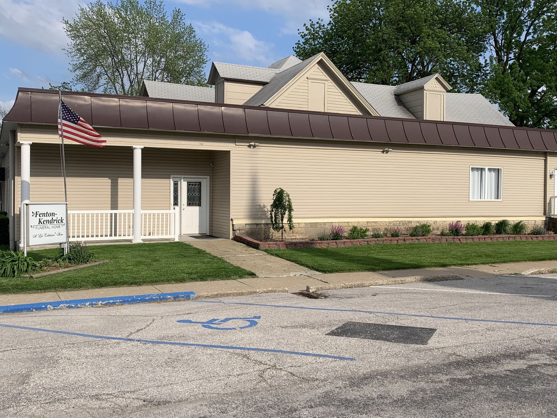 Small beige building with a porch, flag, ramp, and parking lot under a blue sky