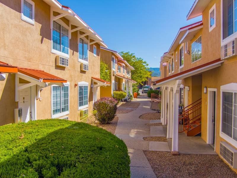 Exterior view of a tan, two-story apartment complex with walkways and landscaping at Santa Fe Suites in Santa Fe, NM.