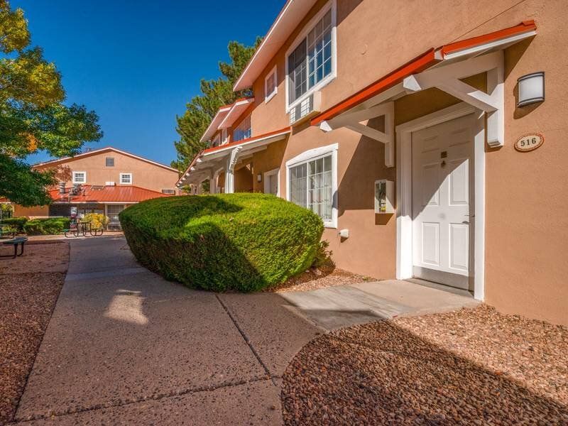 Exterior view of an apartment community building entrance with a white door, walkway, and trimmed hedges at Santa Fe Suites in Santa Fe, NM.