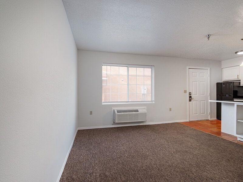 Living room with carpet, a window, and an open kitchen in an apartment unit at Santa Fe Suites in Santa Fe, NM.