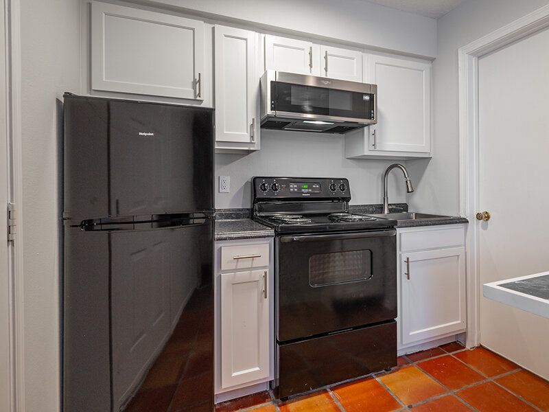 Kitchen in an apartment with a black refrigerator, stove, microwave, white cabinets, and dark countertops at Santa Fe Suites in Santa Fe, NM.