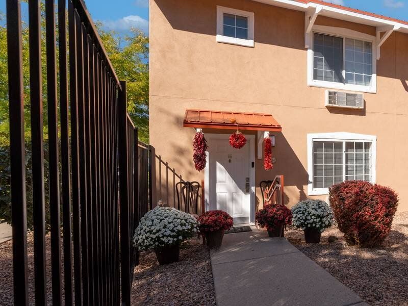 Front entrance of an apartment building with a small red awning, potted flowers, and a metal fence at Santa Fe Suites in Santa Fe, NM.