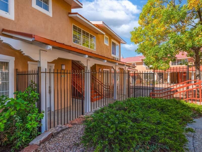 Exterior view of a beige apartment building with stairs, a black metal fence, and landscaped shrubs at Santa Fe Suites in Santa Fe, NM.