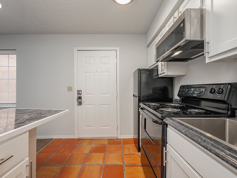 Kitchen in an apartment with white cabinets, black appliances, microwave, and terracotta tile floor at Santa Fe Suites in Santa Fe, NM.