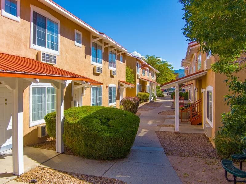Exterior view of a sunny apartment community with tan stucco buildings and a pedestrian walkway at Santa Fe Suites in Santa Fe, NM.