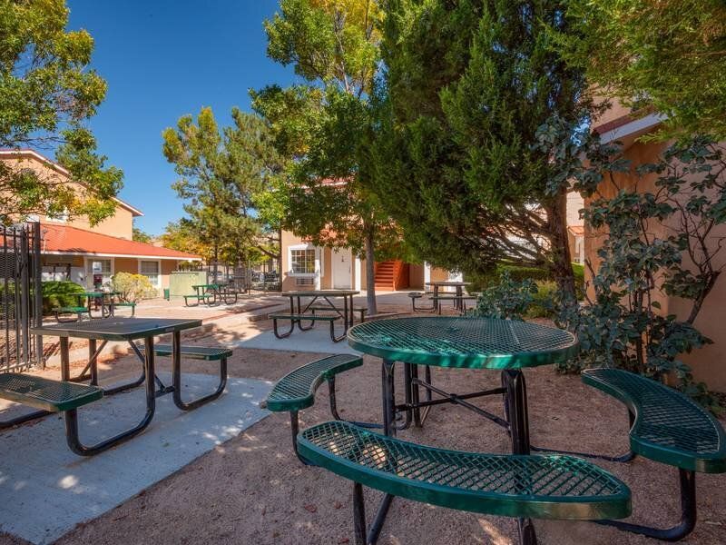 Outdoor communal courtyard with green metal picnic tables, benches, and trees at Santa Fe Suites in Santa Fe, NM.