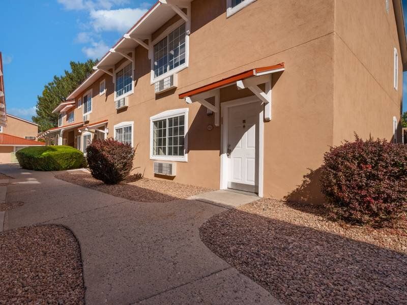 Exterior view of a tan apartment building with white-framed windows, front door, and desert landscaping at Santa Fe Suites in Santa Fe, NM.