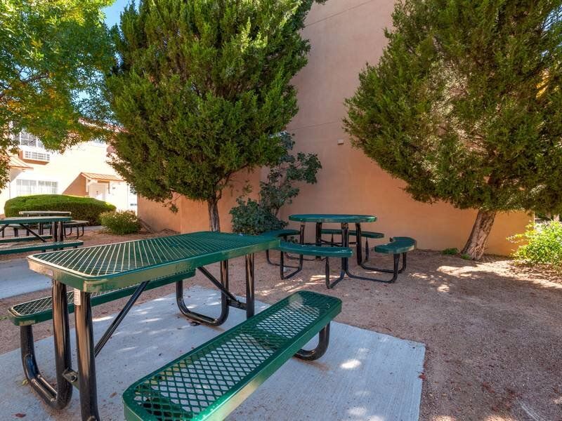 Outdoor communal picnic area with green metal tables and benches under trees beside a beige building at Santa Fe Suites in Santa Fe, NM.