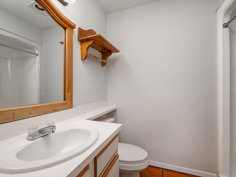 Bathroom in an apartment with a white sink, wooden-framed mirror, shelf, toilet, and shower at Santa Fe Suites in Santa Fe, NM.