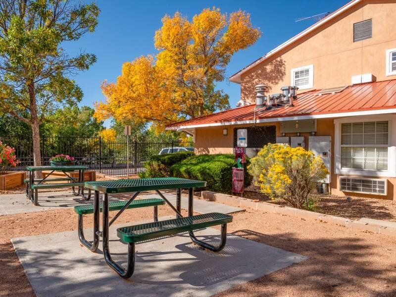 Outdoor community seating area with green picnic tables in a courtyard beside an orange apartment building at Santa Fe Suites in Santa Fe, NM.